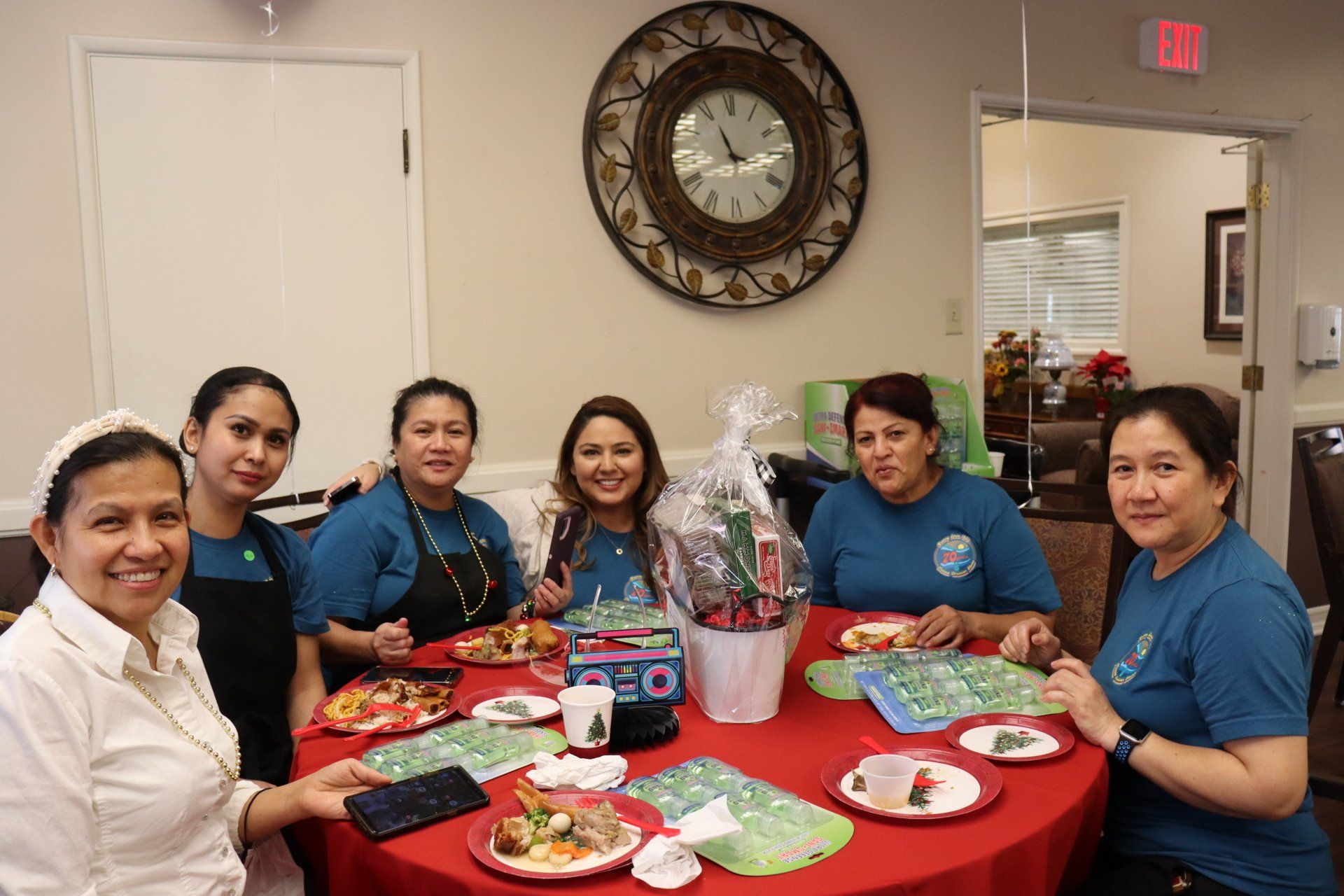 Group of women smiling around a red table, eating and celebrating. Indoor setting with clock on wall.