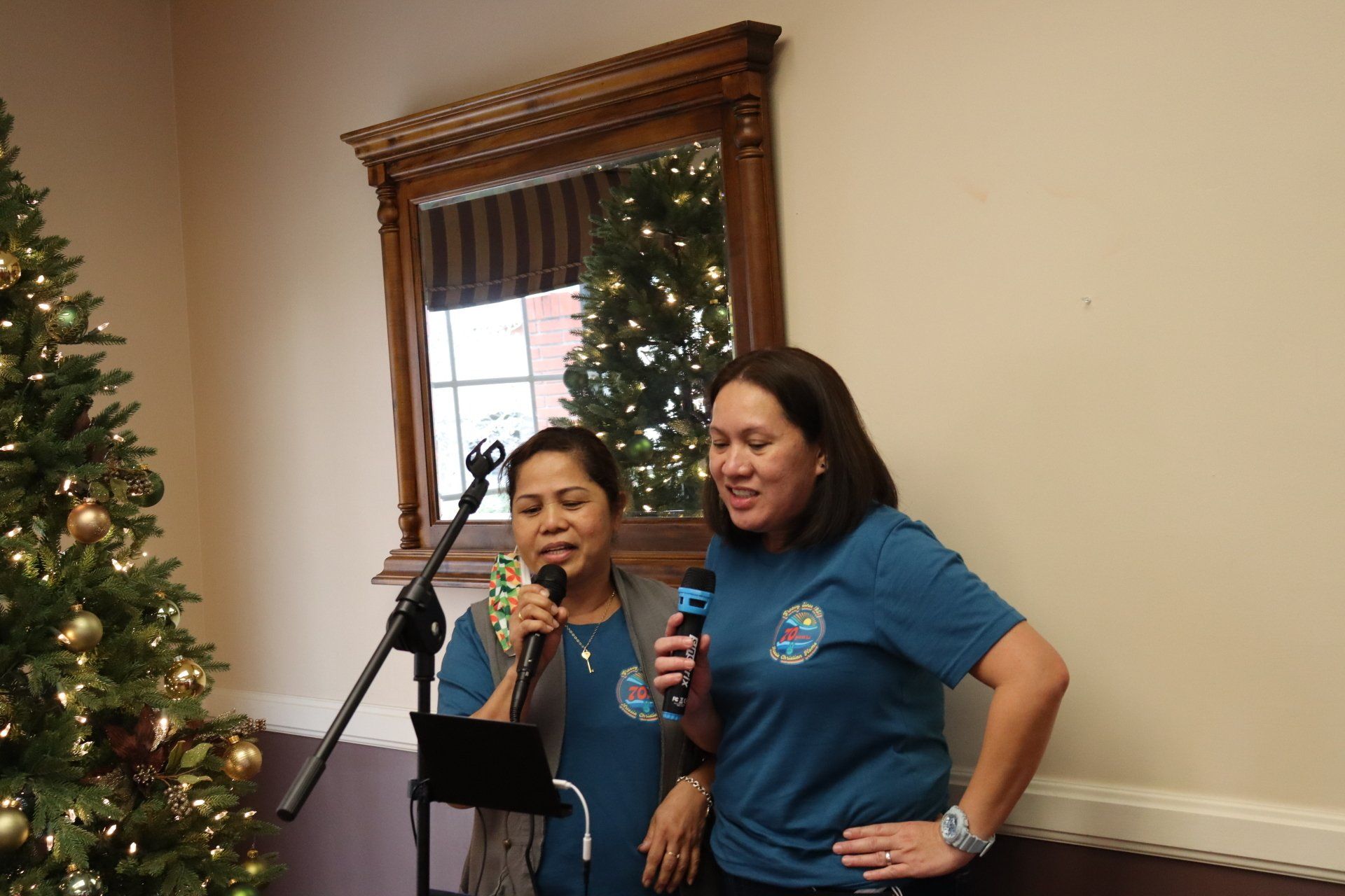 Two women singing karaoke with microphones next to a Christmas tree and mirror.