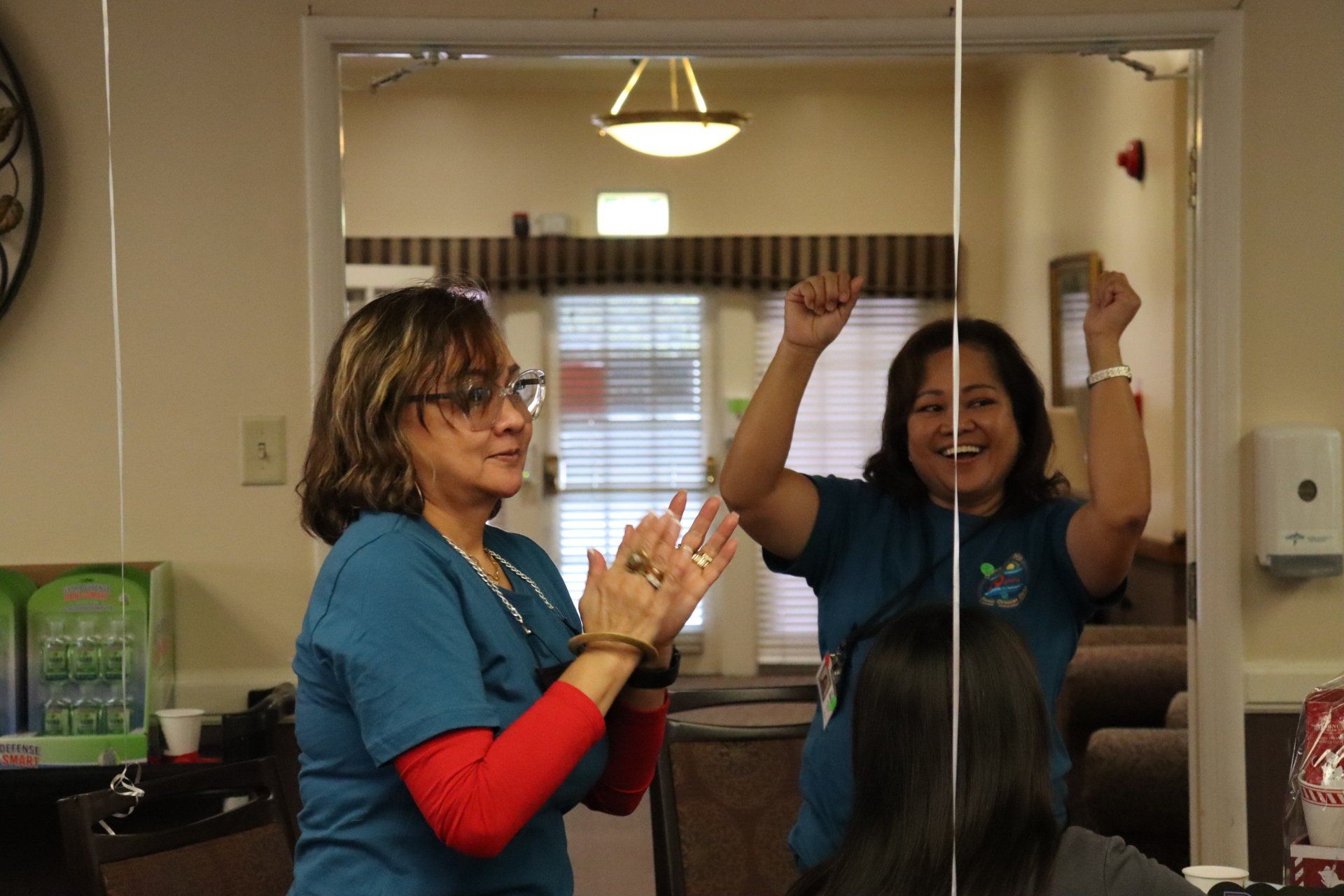 Two women in teal shirts celebrate, clapping and raising arms in an interior setting.