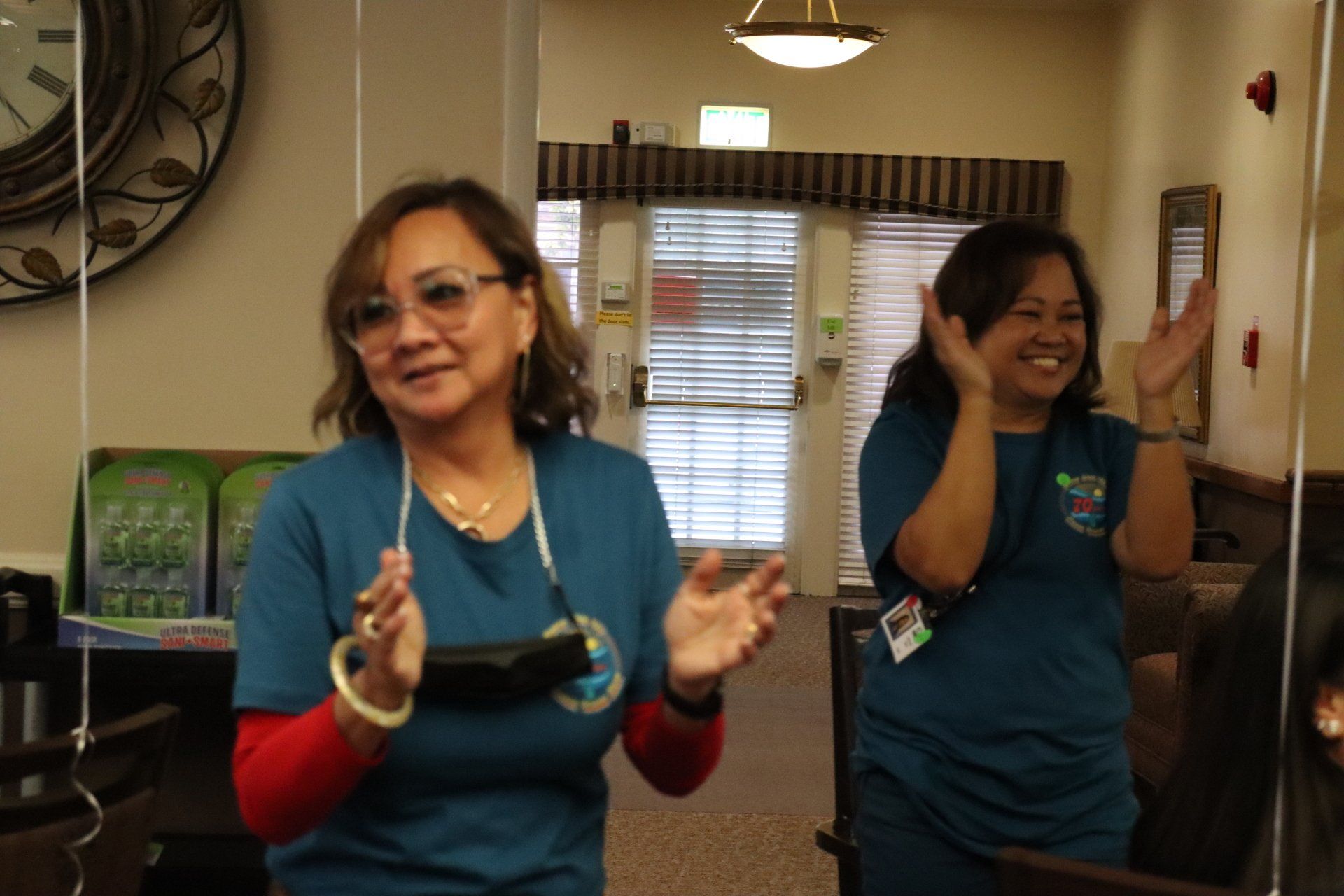Two women in teal shirts clapping in a brightly lit interior hallway.
