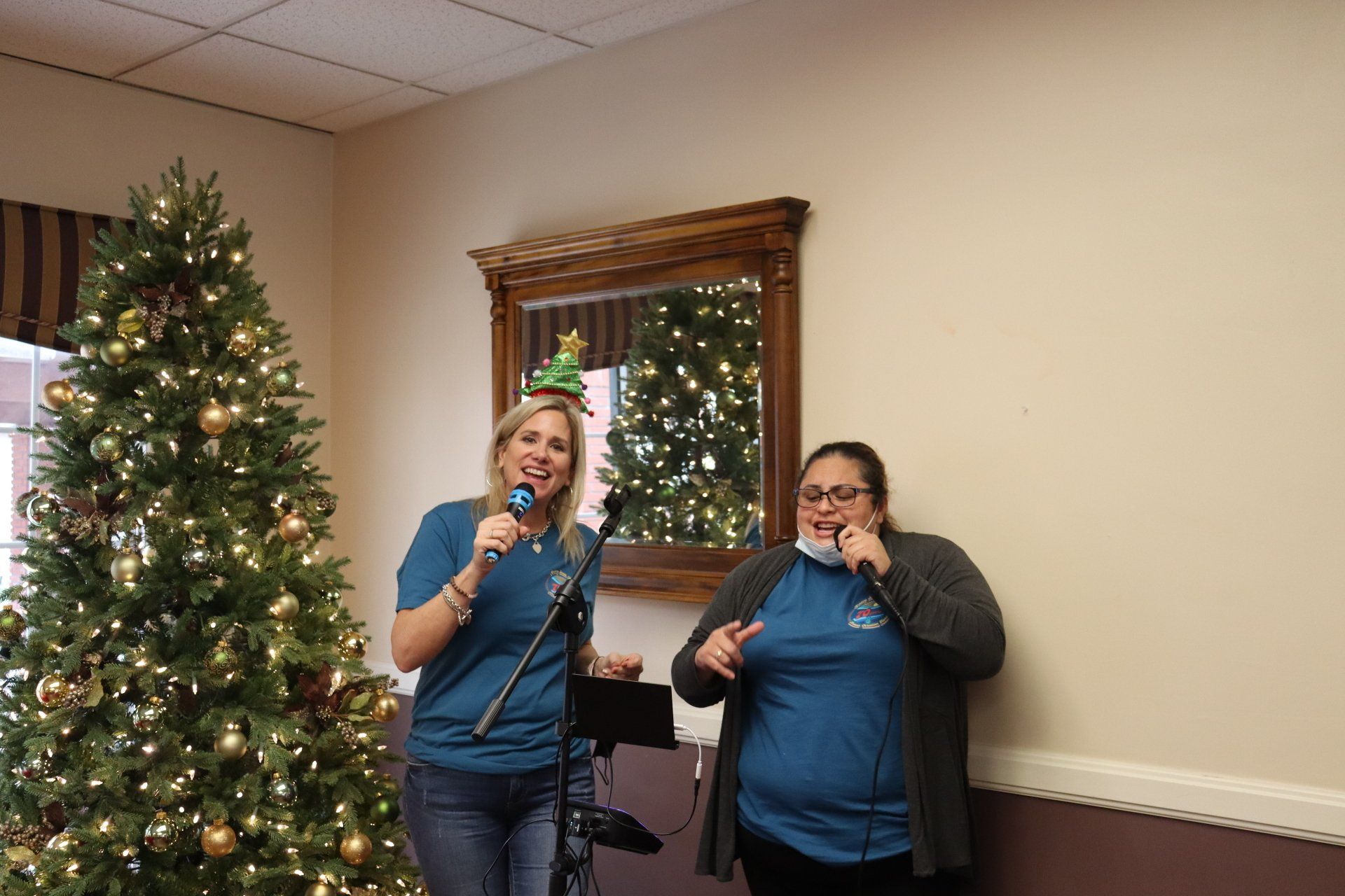 Two women singing into microphones near a Christmas tree and a mirror in a decorated room.