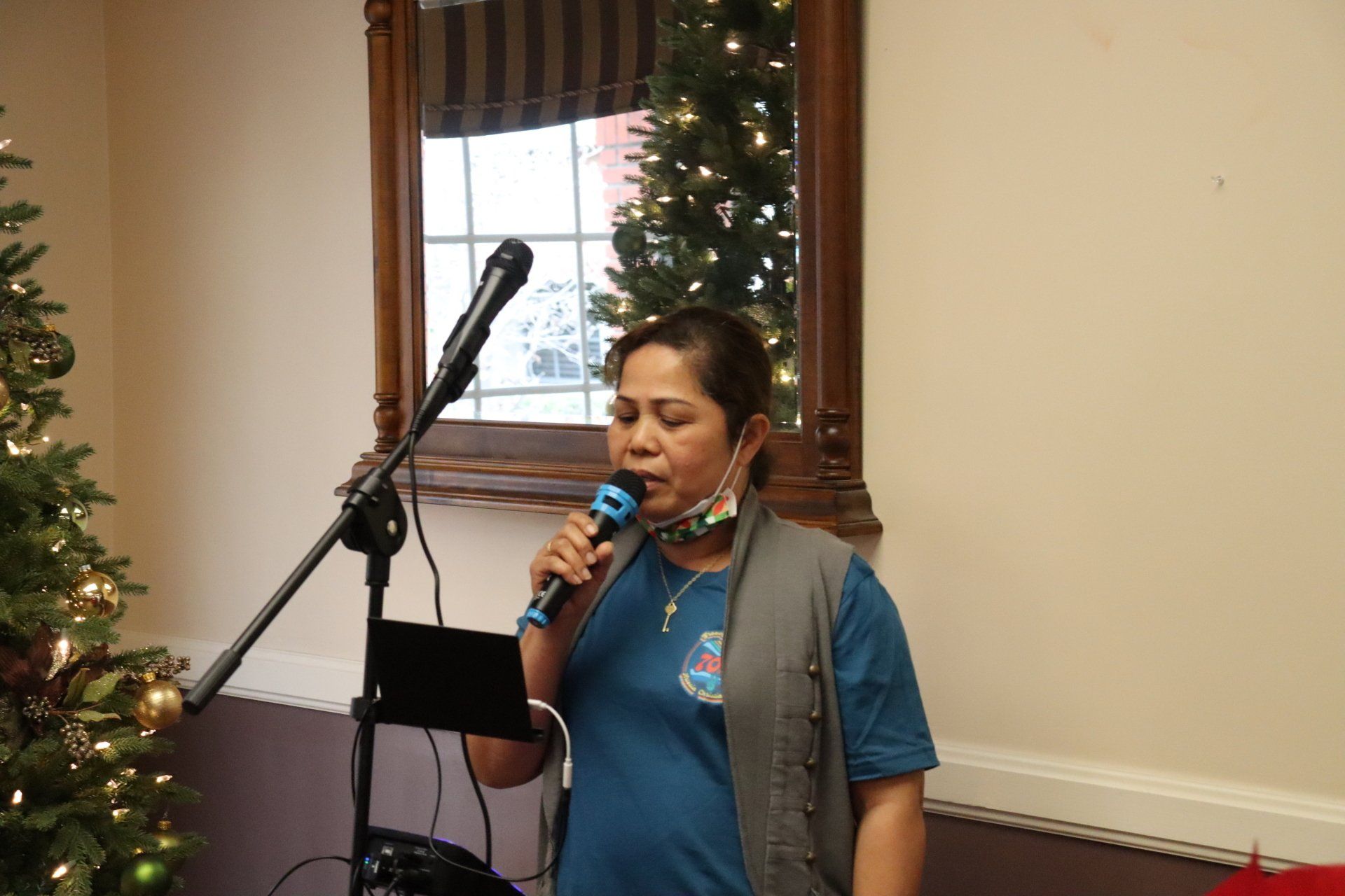 Woman singing into a microphone, standing in front of a Christmas tree and a mirror.