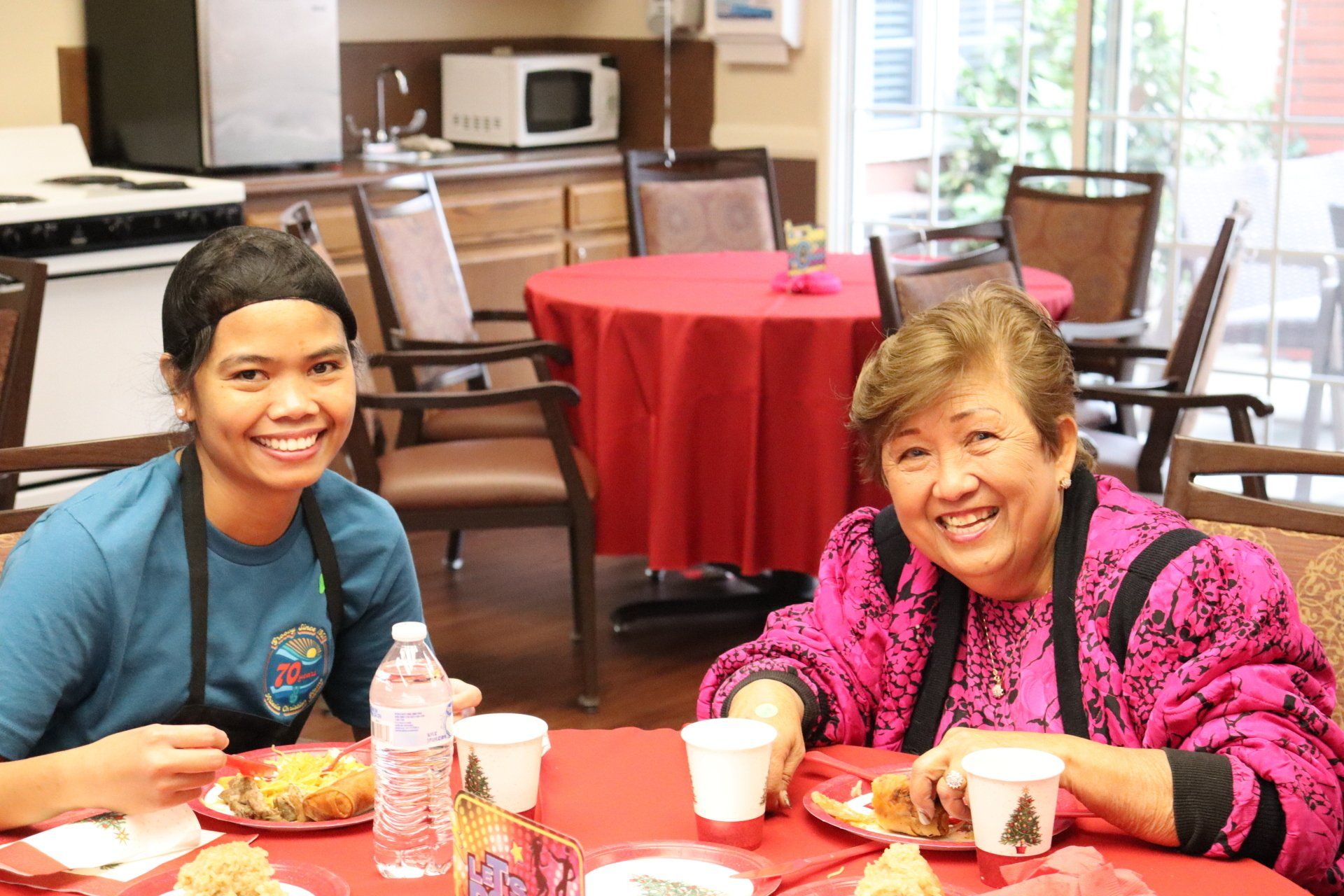 Two smiling women seated at a red table in a dining area; one is wearing a blue shirt, the other, a pink blouse.