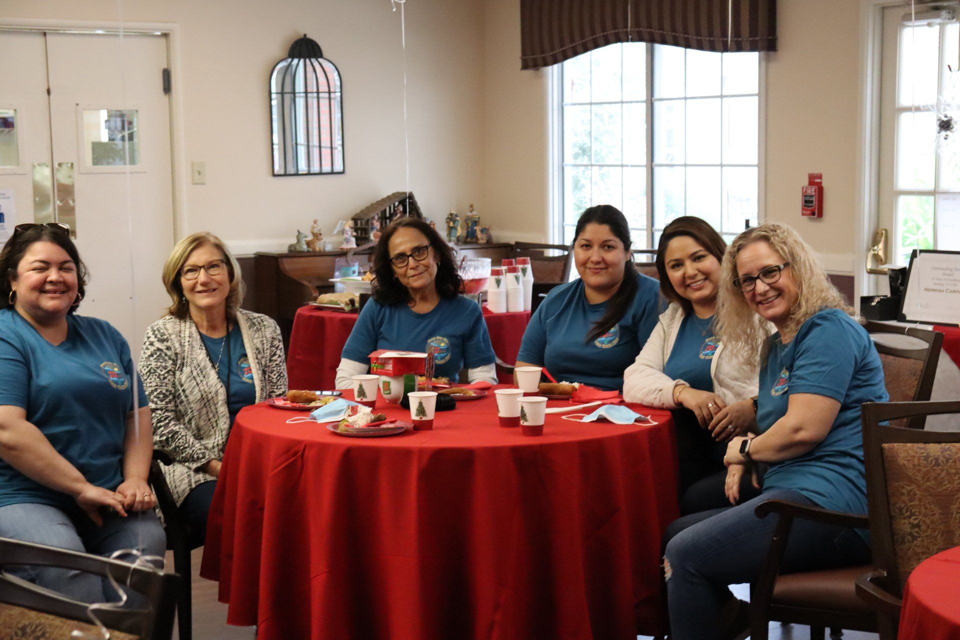 Six women sit at a round table with a red tablecloth, smiling. They are in a room with a window and decorations.