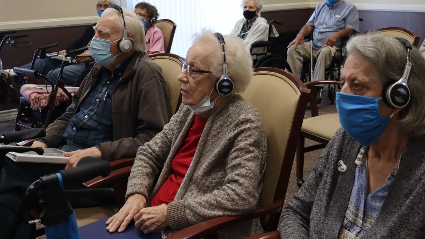 Senior citizens wearing headphones and masks sit in chairs, attending a group event indoors.