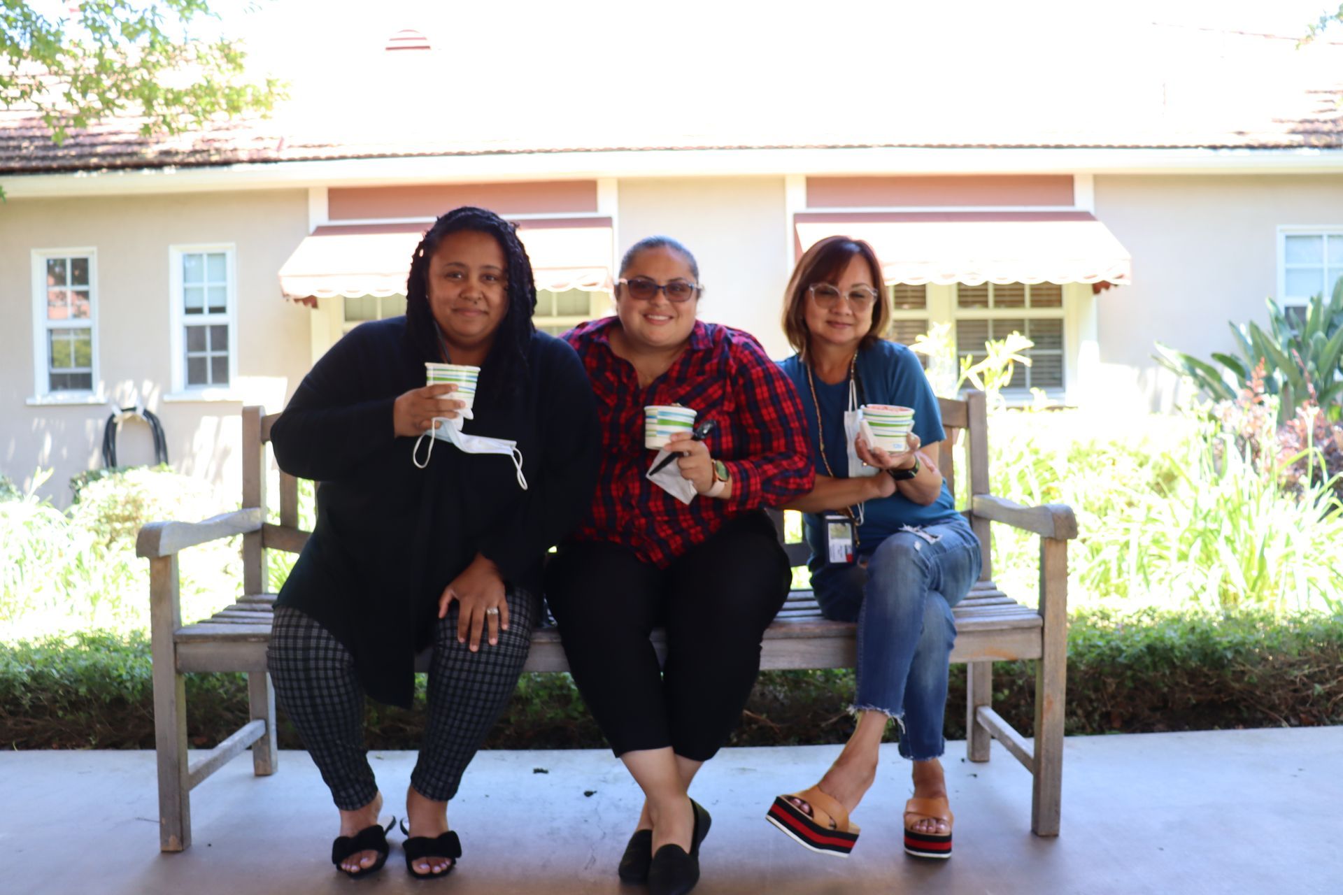 Three women sitting on a bench, holding cups, smiling. They are outside in front of a building.
