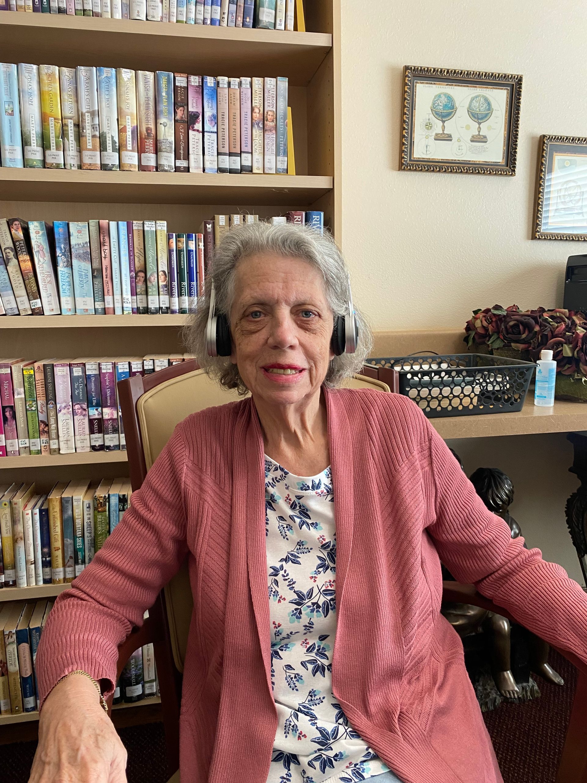 Elderly woman in pink cardigan and headphones sits in a library.