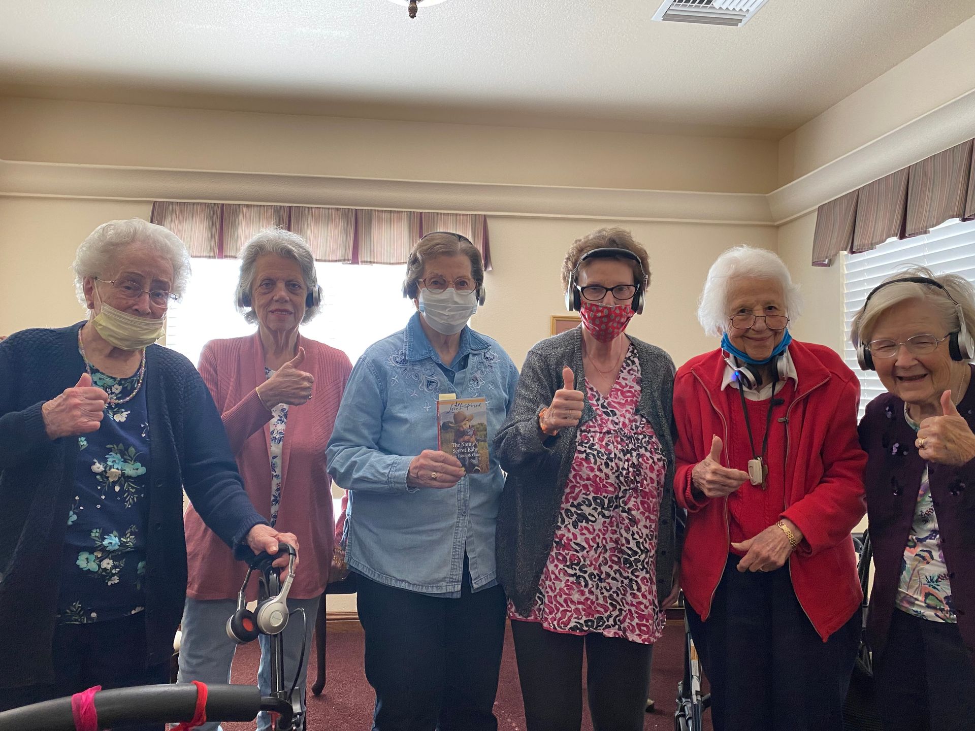 Six senior women wearing masks and headphones give thumbs up, indoors.