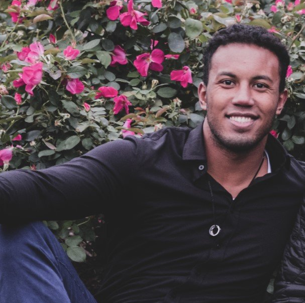 Man smiling outdoors, near pink roses, wearing a black shirt and necklace.