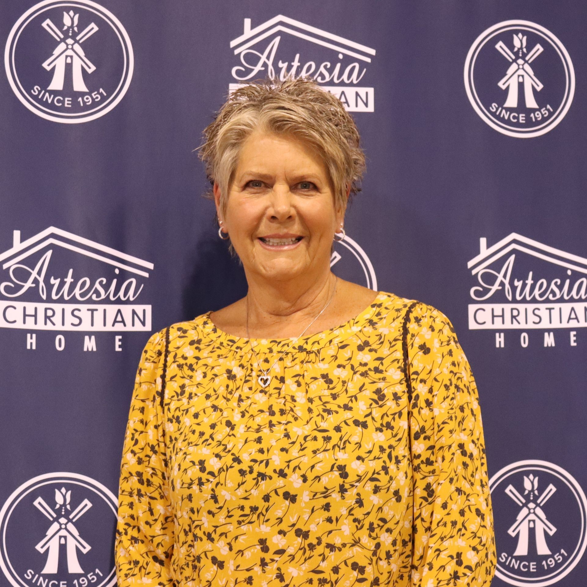 Woman smiling, wearing yellow floral shirt, in front of Artesia Christian Home backdrop.