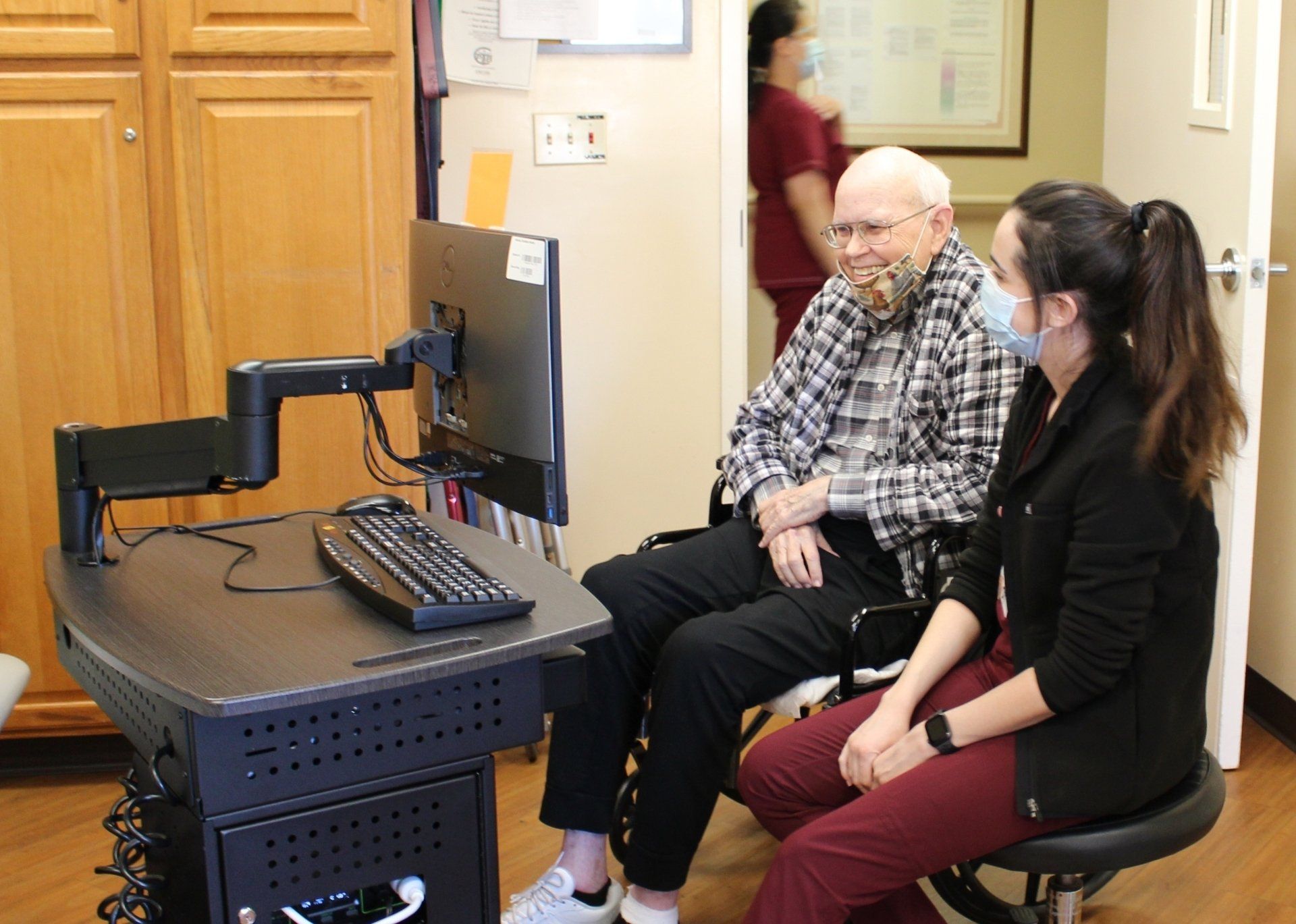 An older man and a woman with masks looking at a computer. The man smiles. They are in an office.