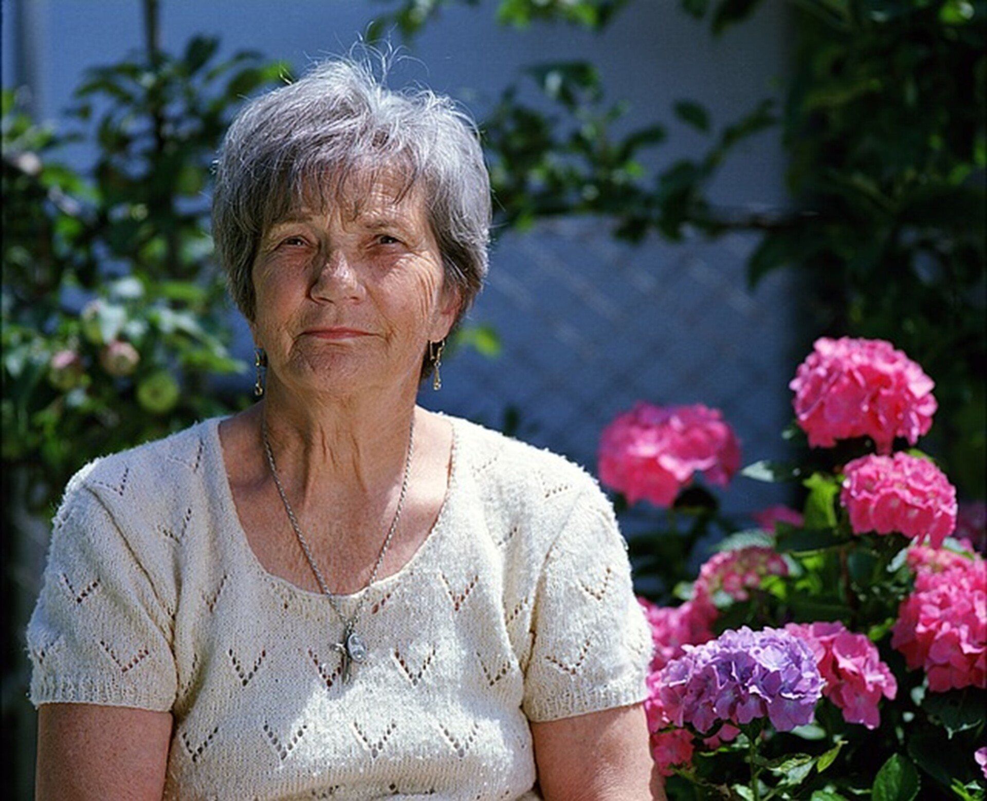 Elderly woman with short gray hair smiles outside near pink and purple hydrangeas.