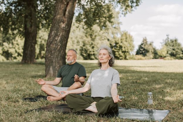 An older couple meditating in a park, sitting cross-legged on a mat, eyes closed, sunny outdoors.