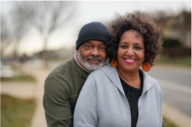 Couple smiling, embracing outdoors. Man wearing black beanie, woman wearing a gray jacket and orange earrings.