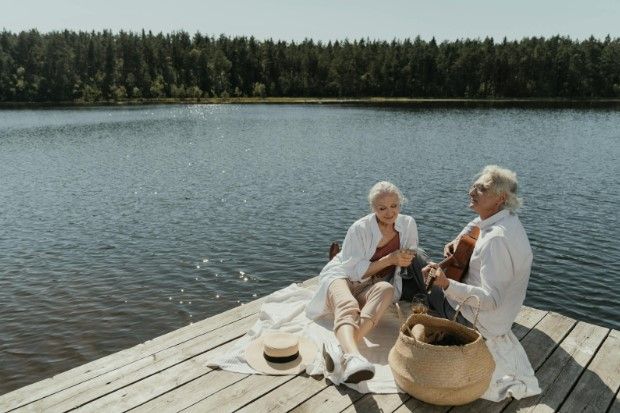Two older adults, enjoying a sunny day at a lake. One plays guitar. Picnic basket, blanket, and trees are in the background.