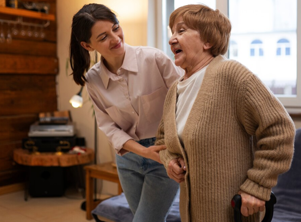Woman helping an elderly woman walk, inside a home. The young woman smiles, holding the older woman's arm.