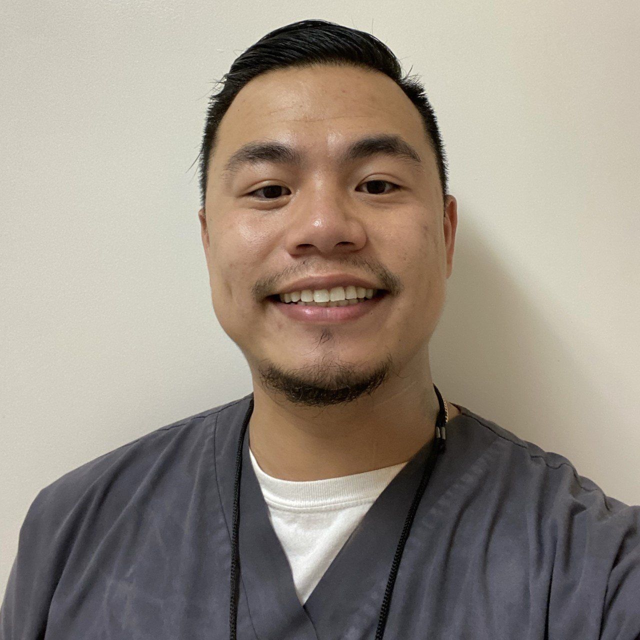 Smiling Asian man in gray scrubs, white shirt, looking at the camera. Neutral background.