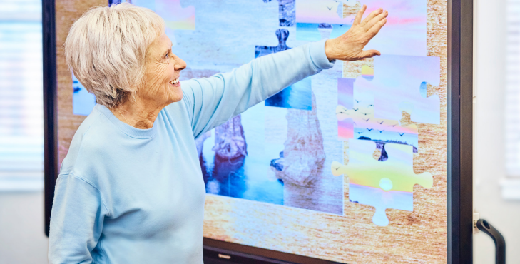 Elderly woman points at a puzzle displayed on a large interactive screen. She wears a blue shirt and smiles.