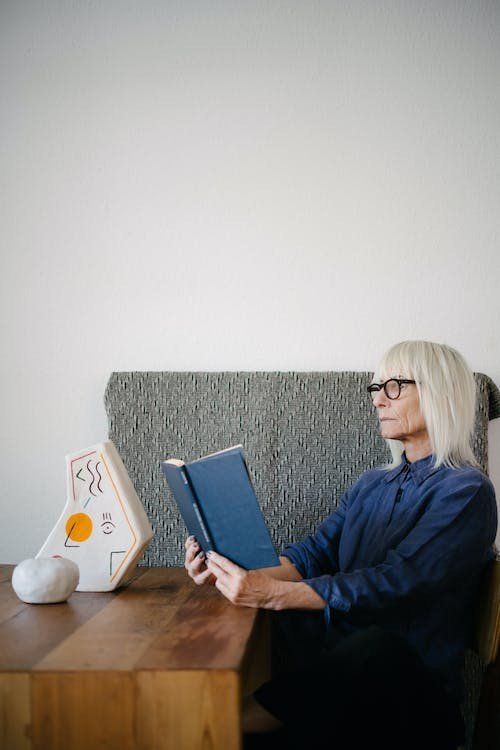 Woman with glasses reading a blue book, sitting near a table with art, against a neutral wall.