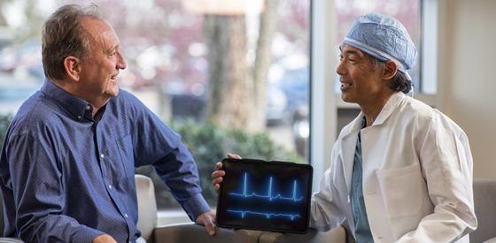 A doctor in scrubs shows an older patient a tablet displaying a heart rhythm. Both are smiling.