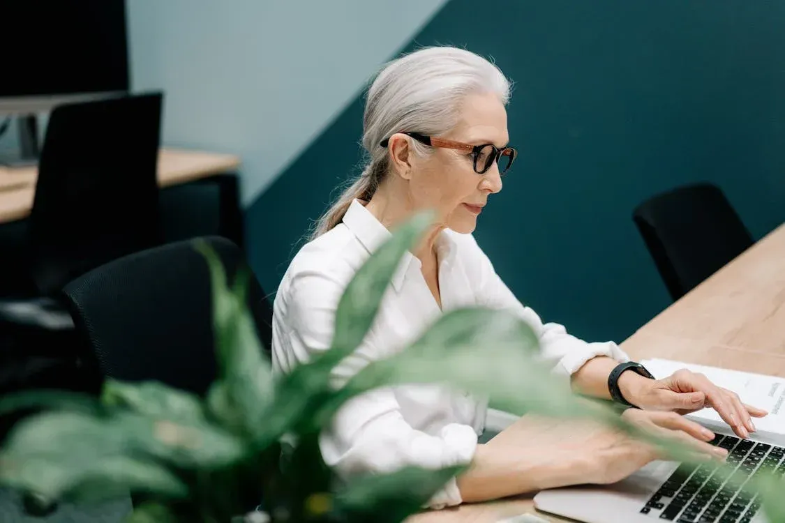 Woman with glasses and gray hair typing on a laptop at a desk in an office.