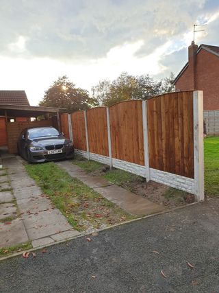 Brown wooden fence with concrete posts bordering a driveway, car parked in a carport.