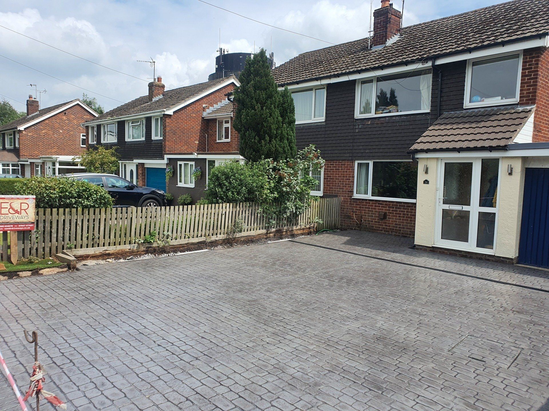 Row of semi-detached houses with driveways and small front gardens, wooden fence, cloudy sky.