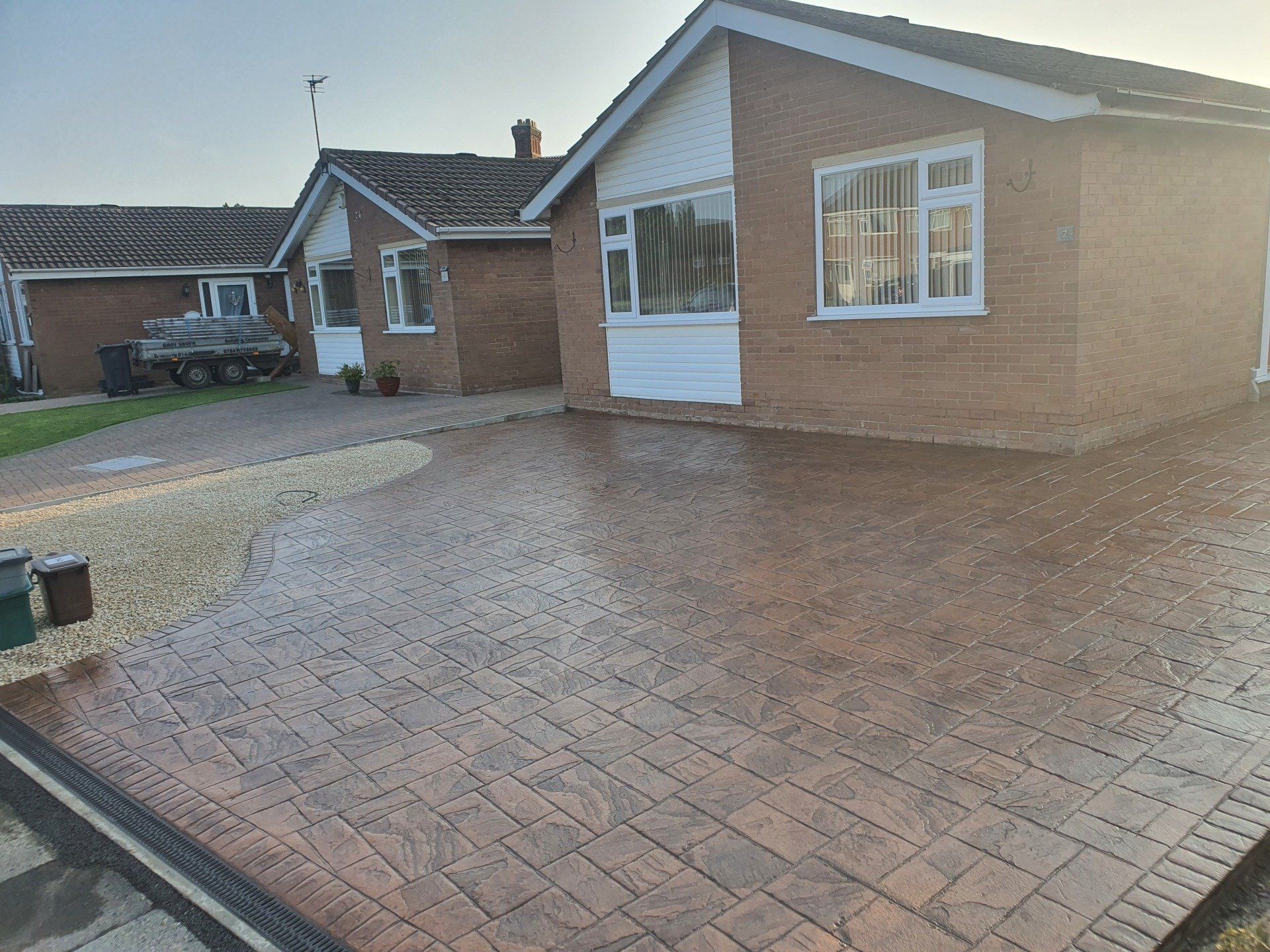 Brick-patterned driveway in front of a brick bungalow with white trim, sunlight glinting off the wet surface.