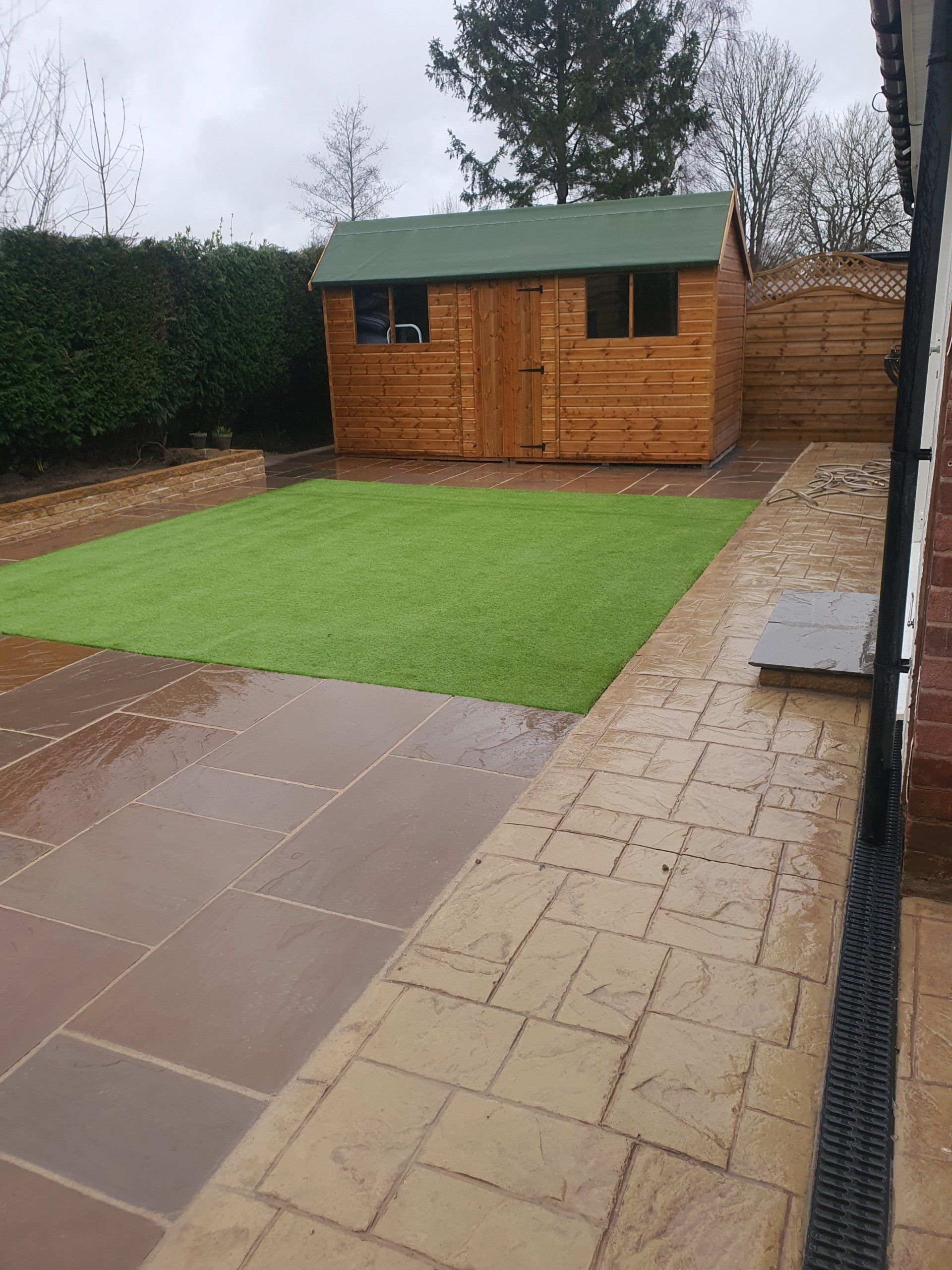 Backyard patio with green turf, brown shed, and brick home exterior.