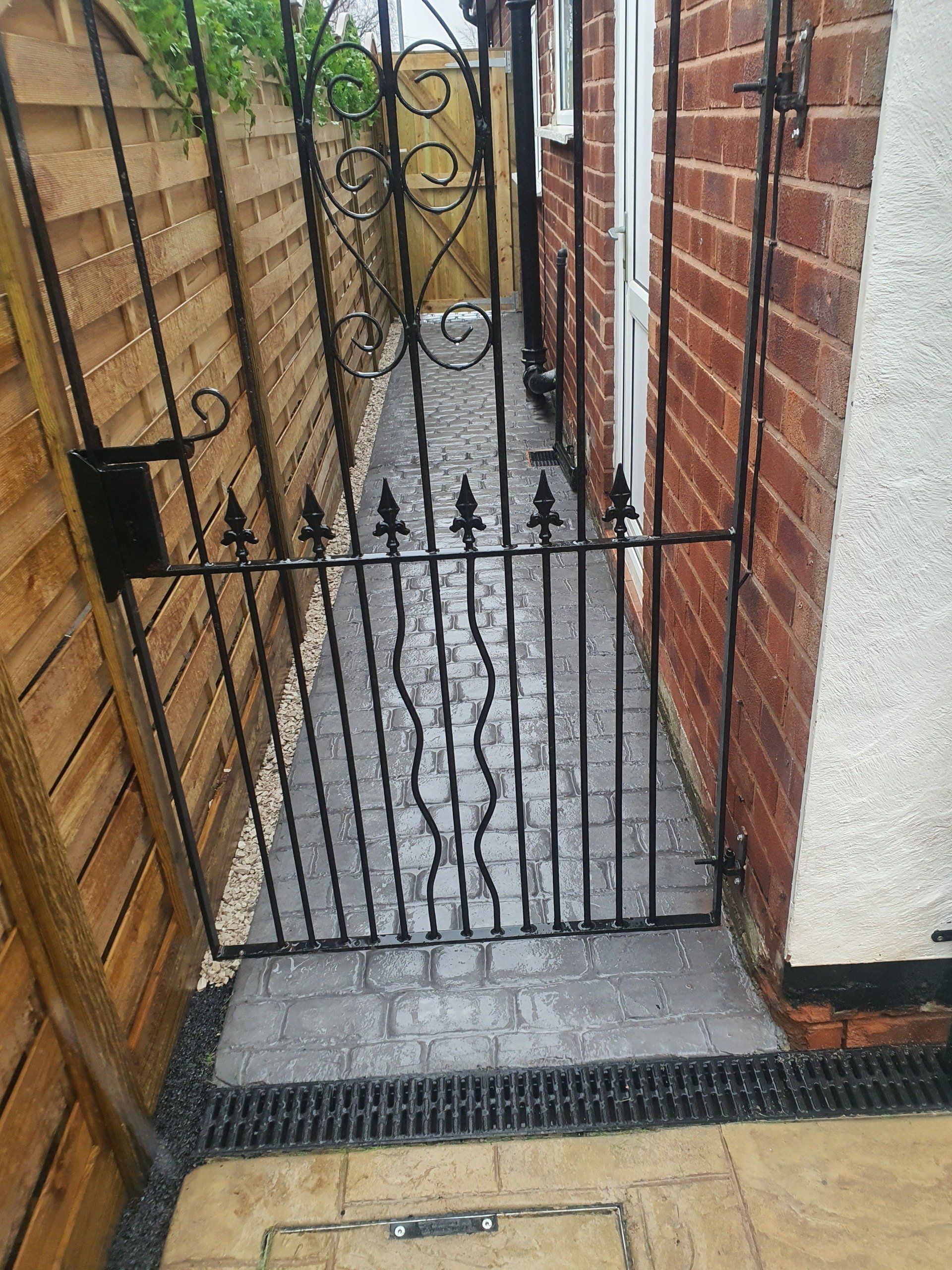 Black wrought iron gate in a narrow alley between a wooden fence and brick wall. Stone path.