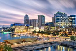 Cityscape at dusk, reflecting in water, with tall modern buildings and a waterfront plaza.