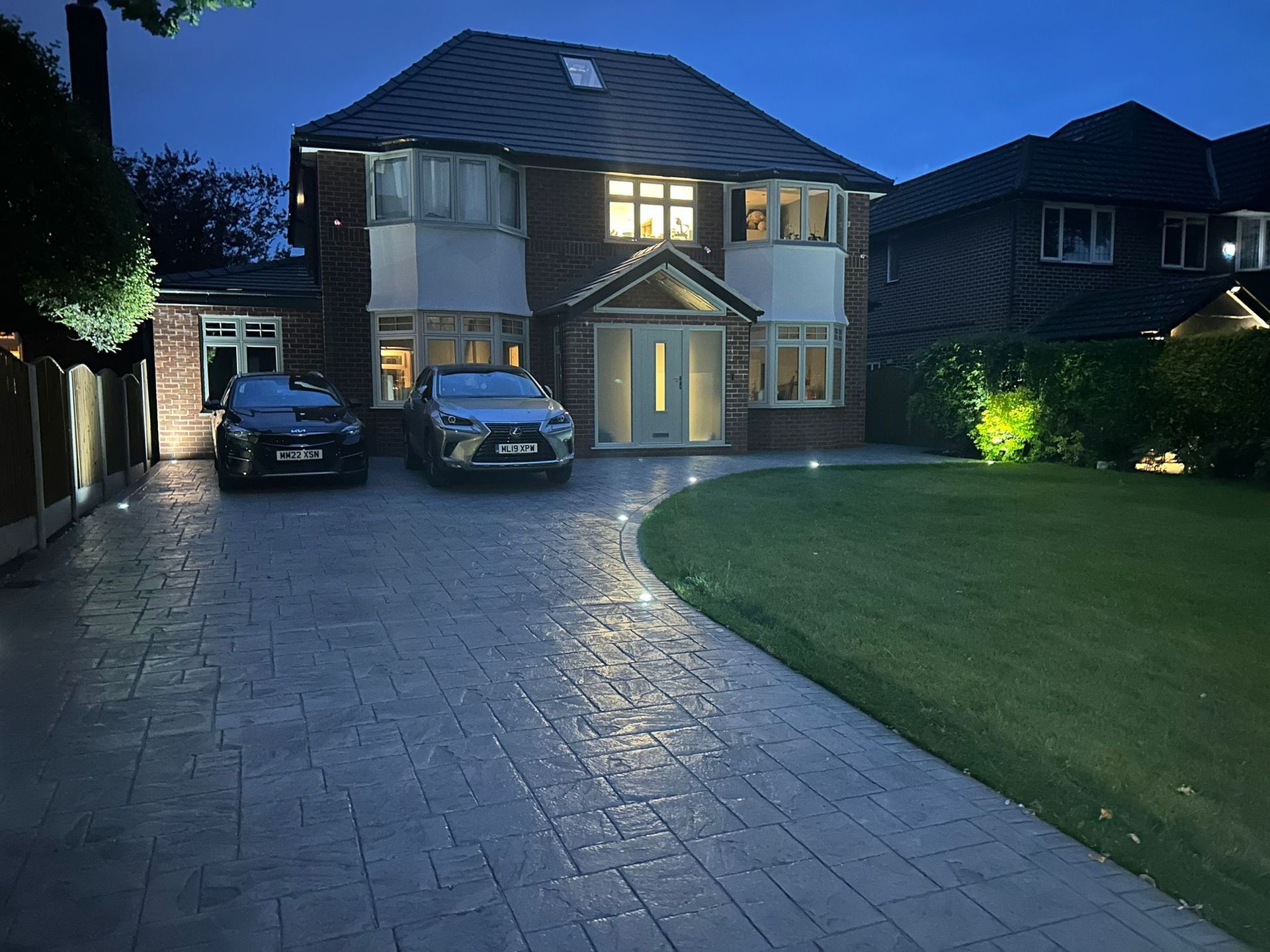 Night view of a two-story house with illuminated driveway and parked cars. Lawn on the right.