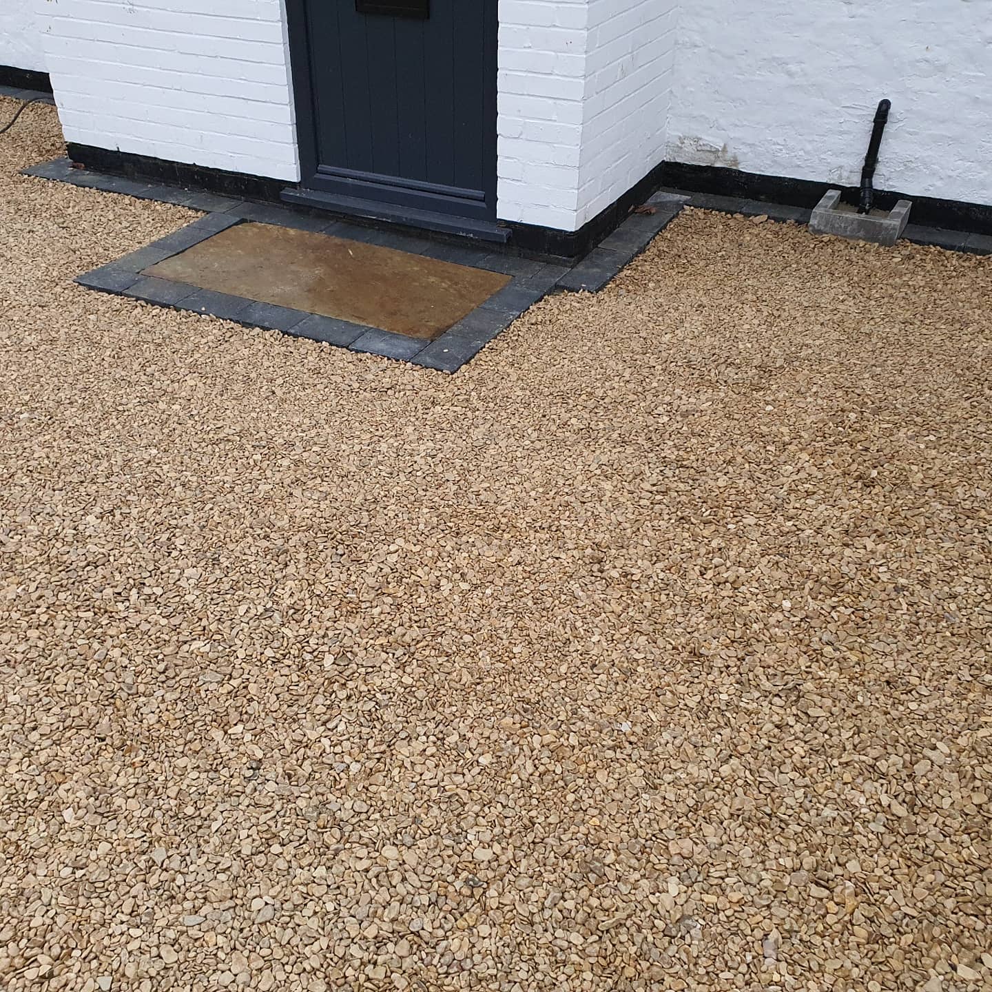 Tan gravel driveway leading to a white brick building with a dark door and doormat.