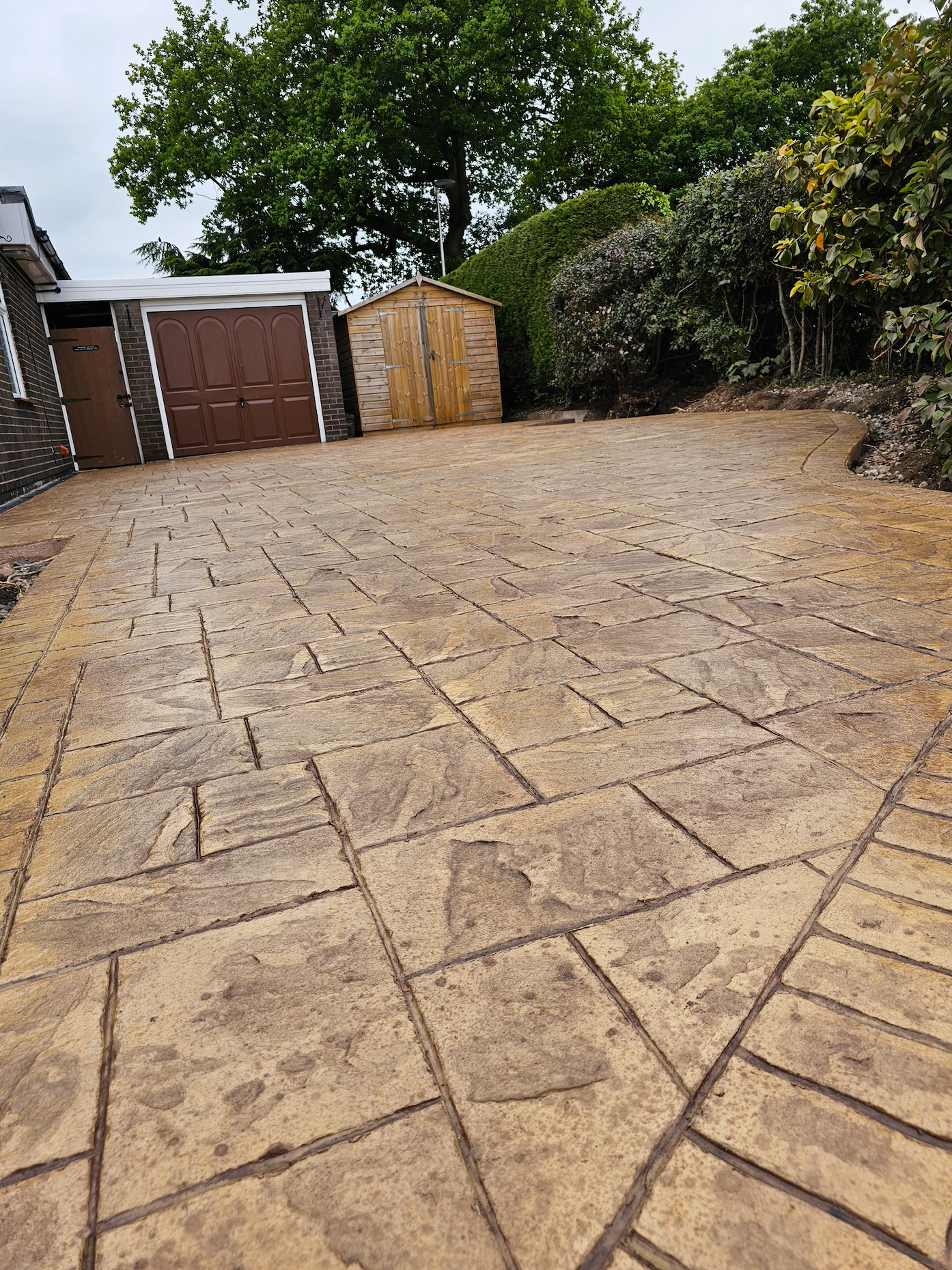 Stamped concrete driveway with brown garage and shed, surrounded by trees.