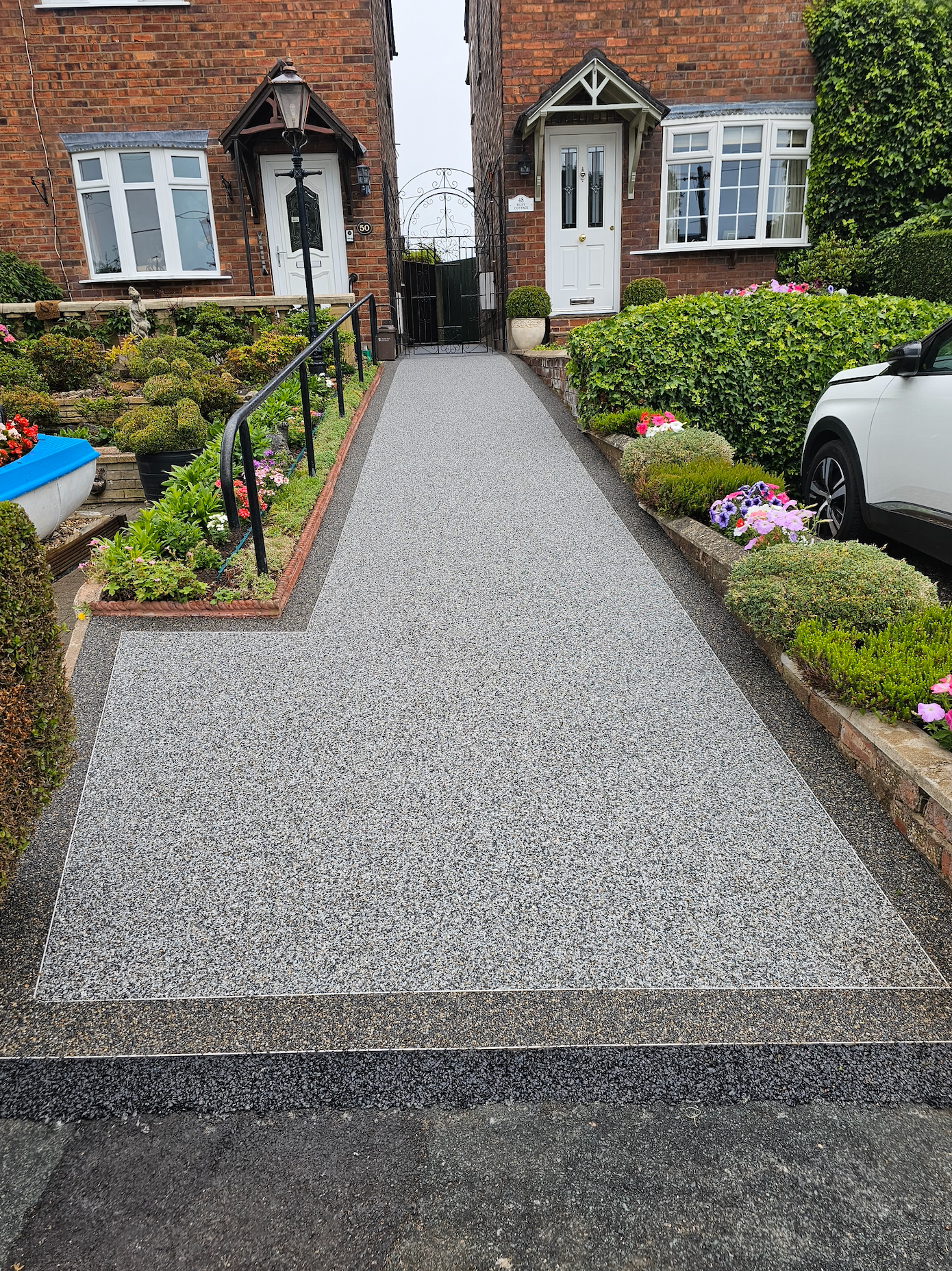 Gravel path between two brick houses with low flowerbeds on either side. A car is parked on the right.