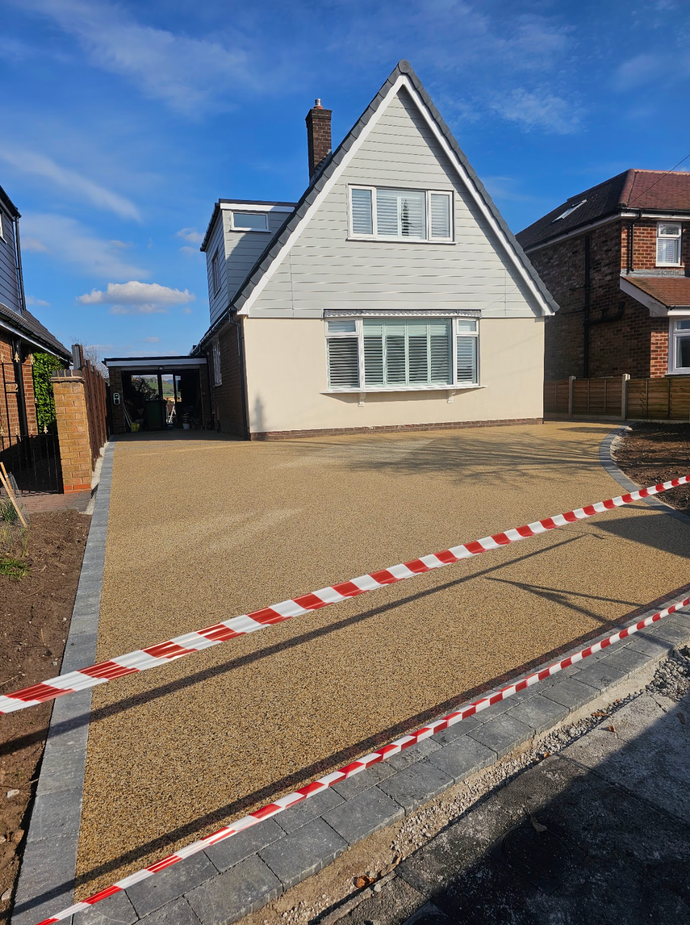 Gravel driveway leading to a two-story house with a grey and beige exterior, bordered by grey paving stones and a red/white striped barrier.