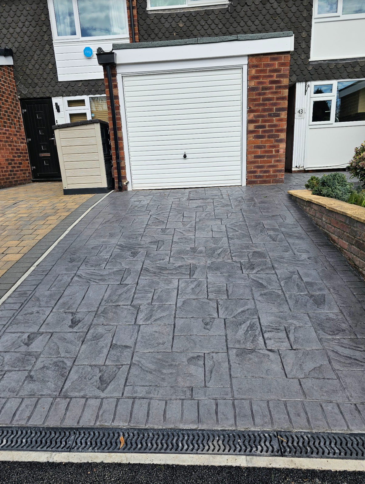 Gray patterned concrete driveway in front of a house with a white garage door. Brick accents and a drainage gutter are present.