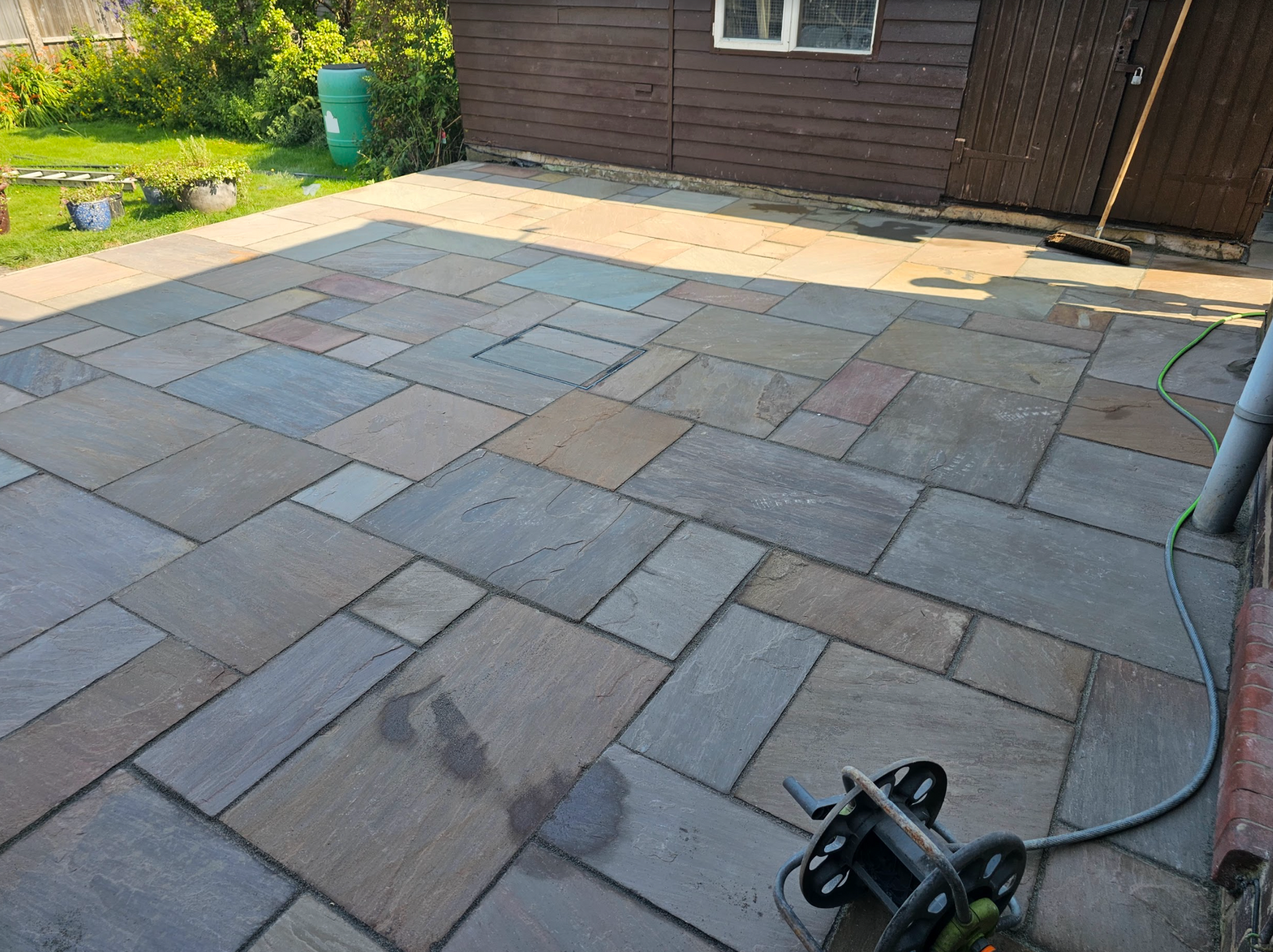 Stone patio with mixed colors, next to a brown wooden shed. Garden hose and pressure washer present.