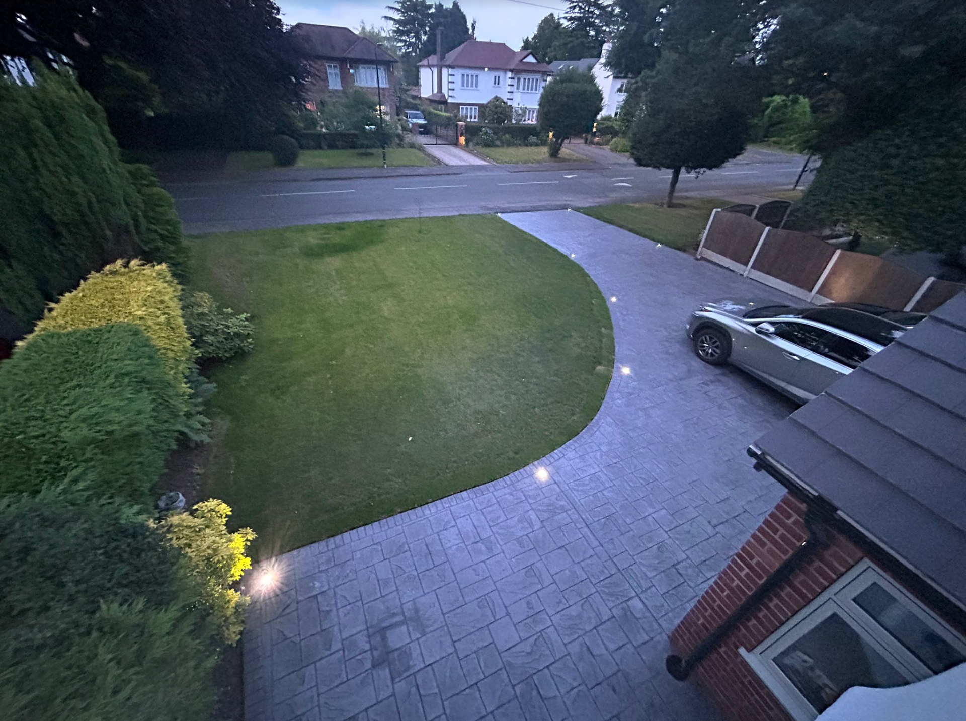 A residential exterior scene at dusk. A paved driveway curves into a lawn, with a car parked.