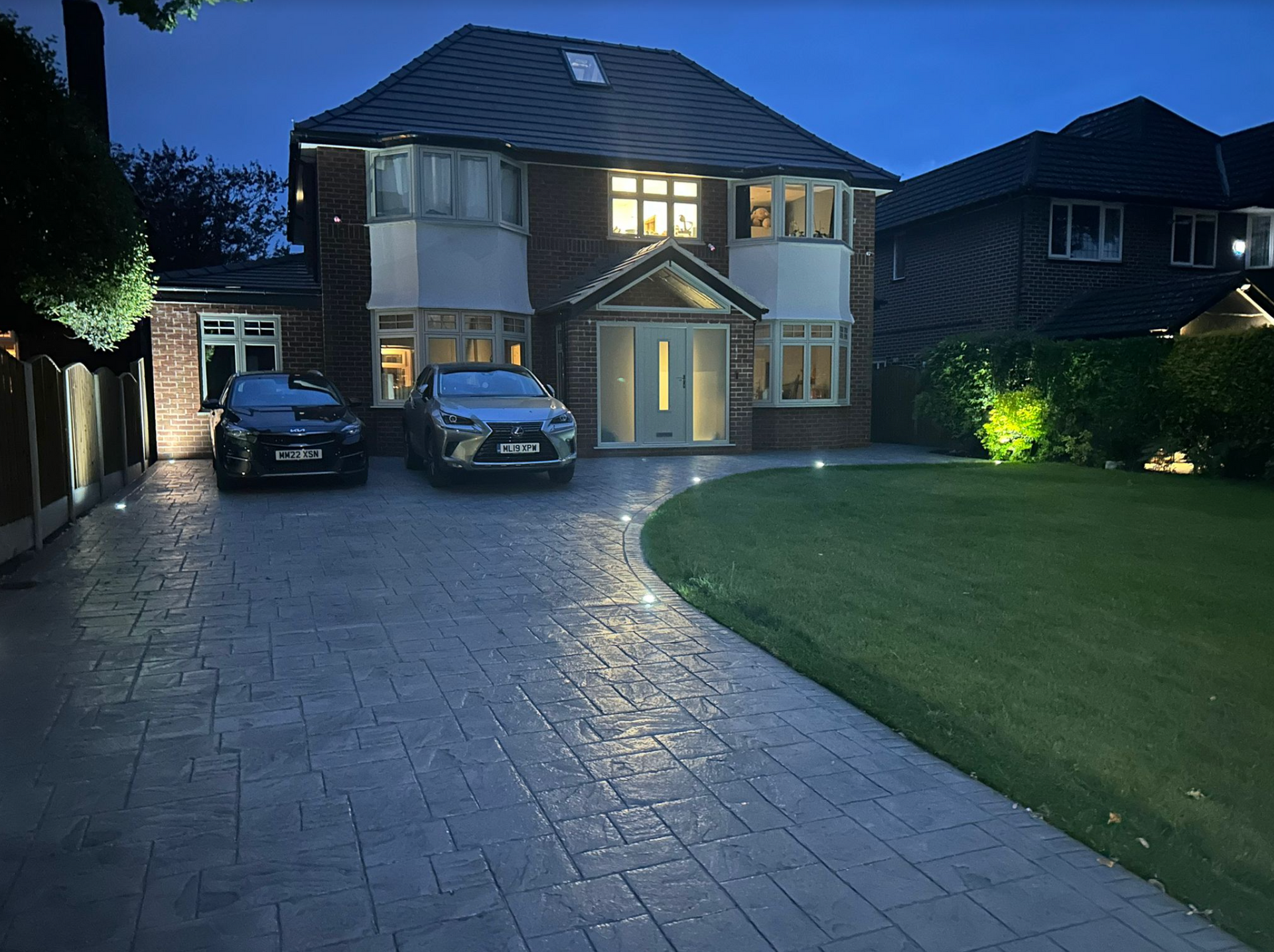 Two-story house with illuminated driveway and two cars parked in front at dusk. Green lawn to the side.