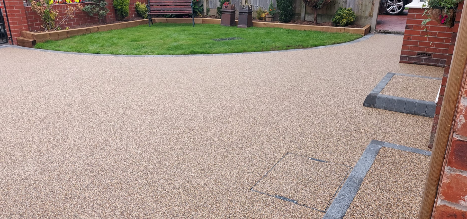 A driveway made of small, tan pebbles curves towards a green lawn with a red brick border.