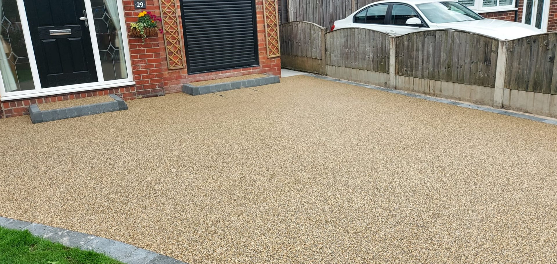 A gravel driveway in front of a brick house, a black door, and a white car parked near a fence.