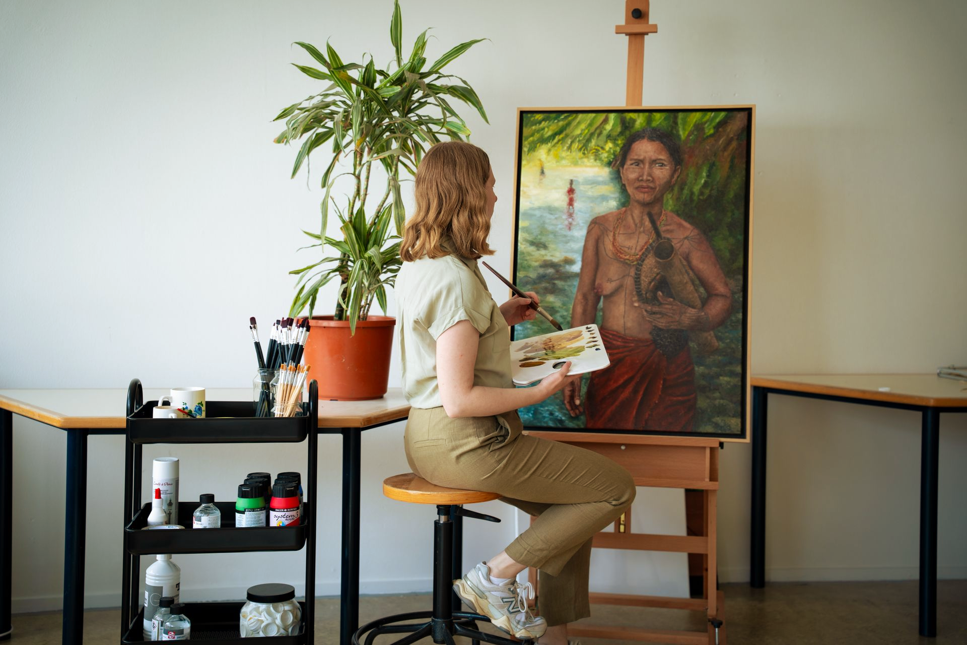 Kunstenaar schildert een zittend figuur op canvas in een atelier met schildersezel, plant en schildersbenodigdheden.