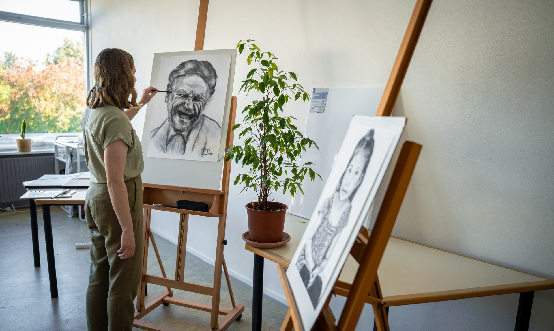 Kunstenaar schetst een portret van een bebaarde man in een lichte studio met schildersezels en een potplant.