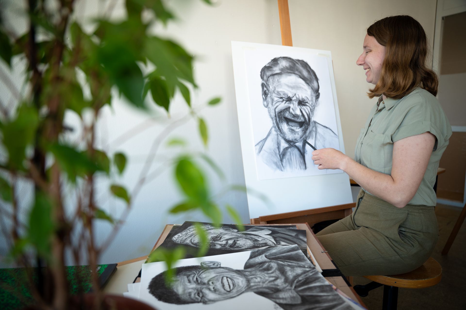 Kunstenaar schetst een portret in houtskool naast een grote tekening op een schildersezel in een lichte studio.