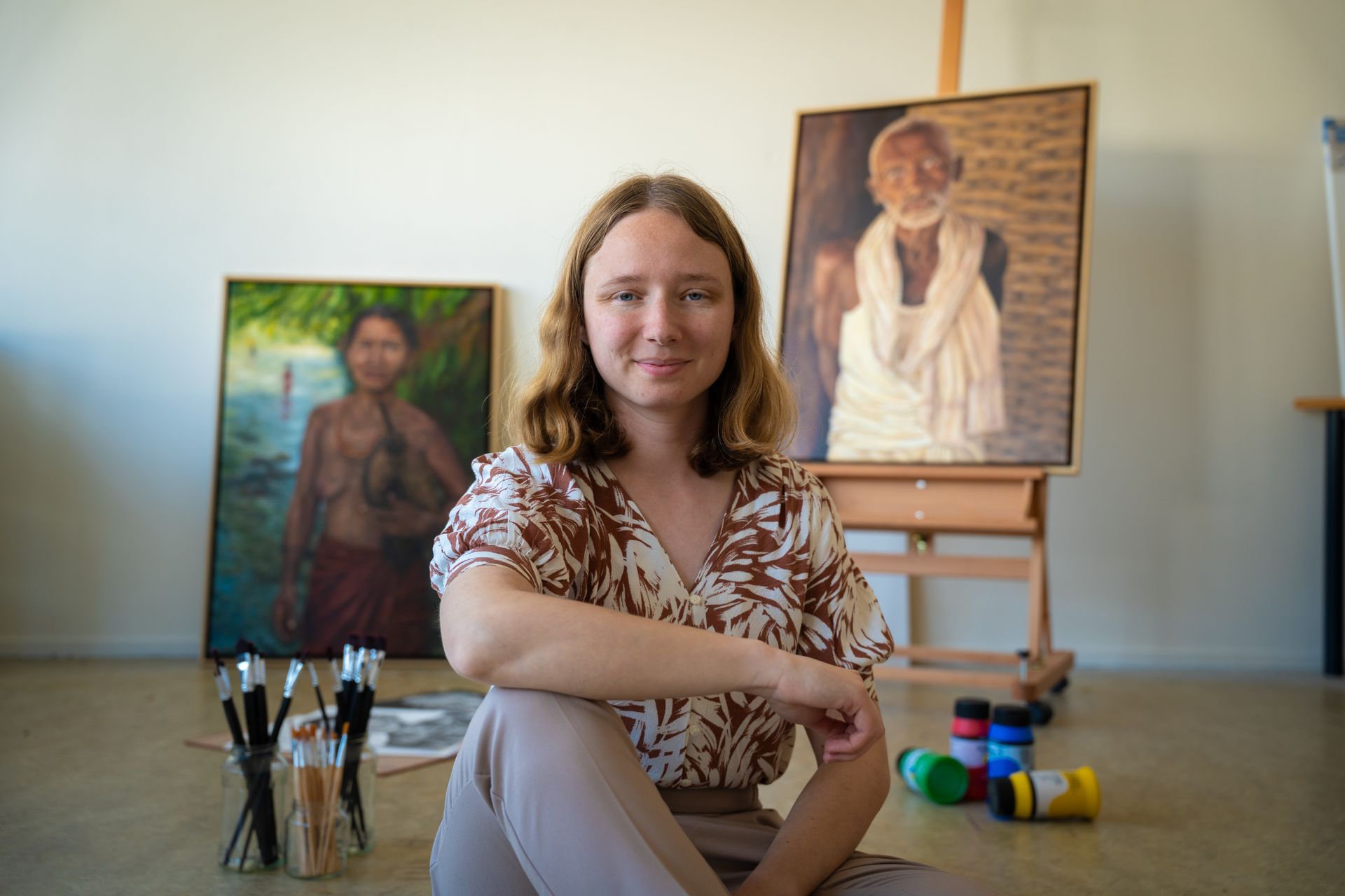 Vrouw zit in een kunststudio met twee portretschilderijen, penselen en schildersbenodigdheden in de buurt.