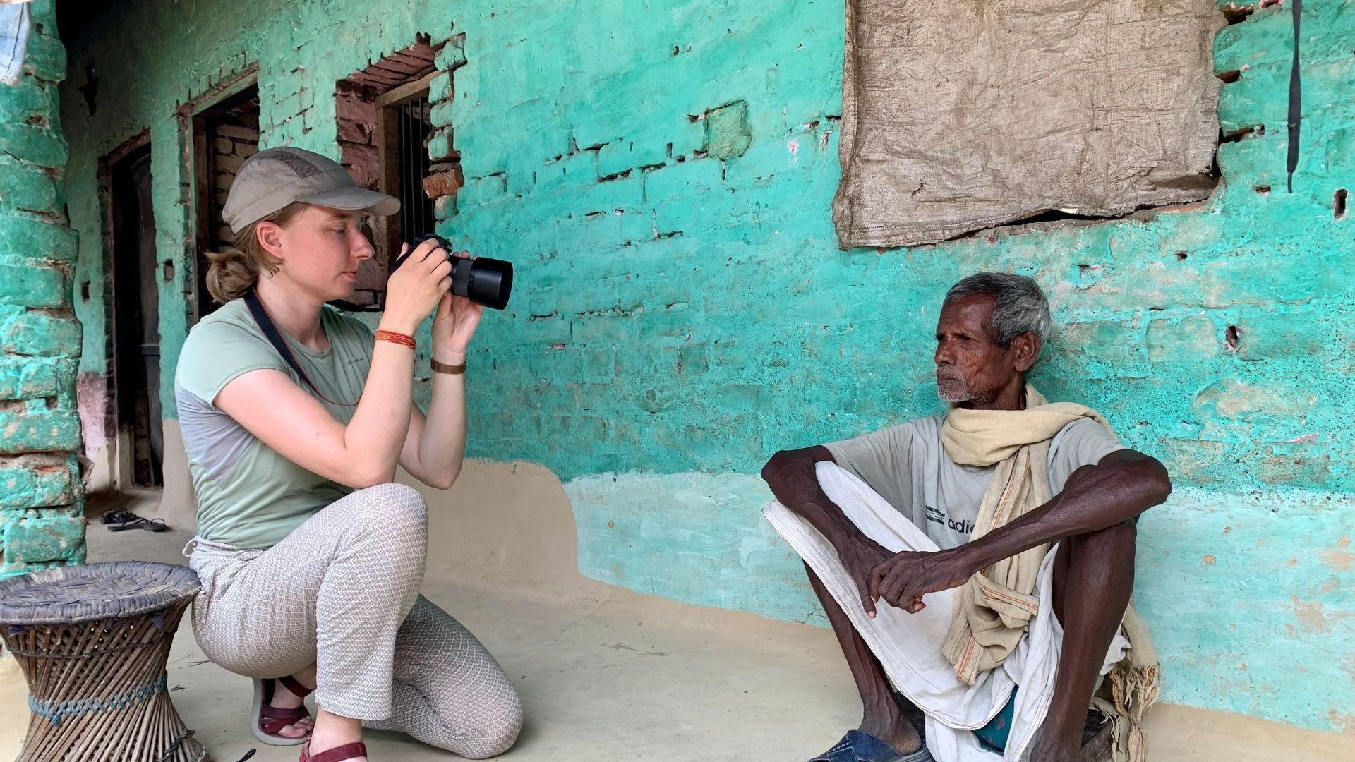 Een vrouw knielt en fotografeert een zittende man tegen een turquoise muur in een rustieke kamer.