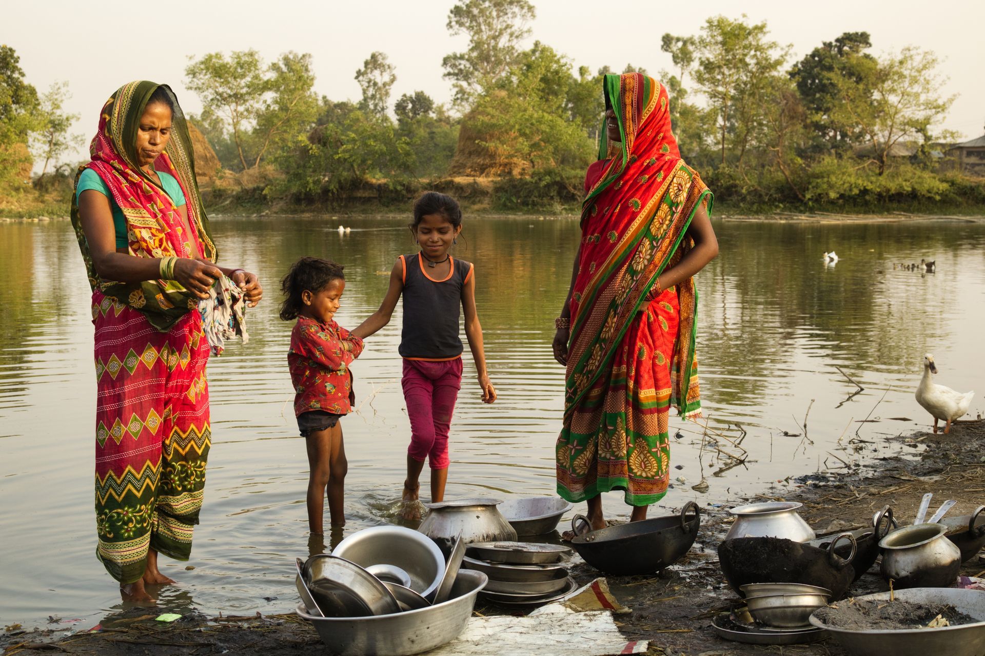 Twee vrouwen en twee kinderen wassen de afwas bij een rivier, met kleurrijke kleren en potten op de voorgrond.