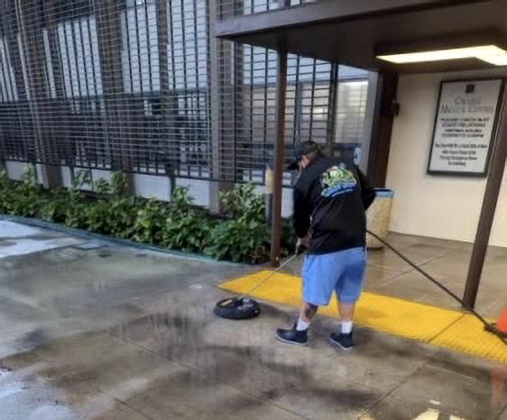 Worker sweeping a wet walkway beside a building entrance with a yellow curb and potted plants.