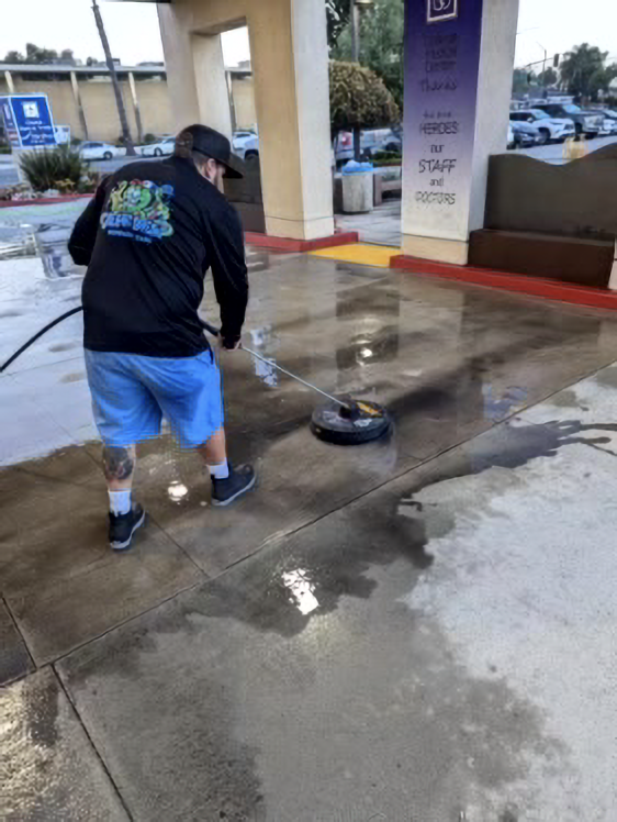Person pressure-washing a wet concrete sidewalk at a gas station, with water spraying across the ground.